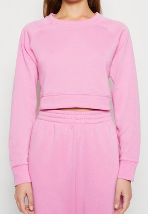 Frau trägt einen langärmeligen, kurzen pinken Pullover und passende hoch taillierte pinke Jogginghosen vor einfachem Hintergrund.