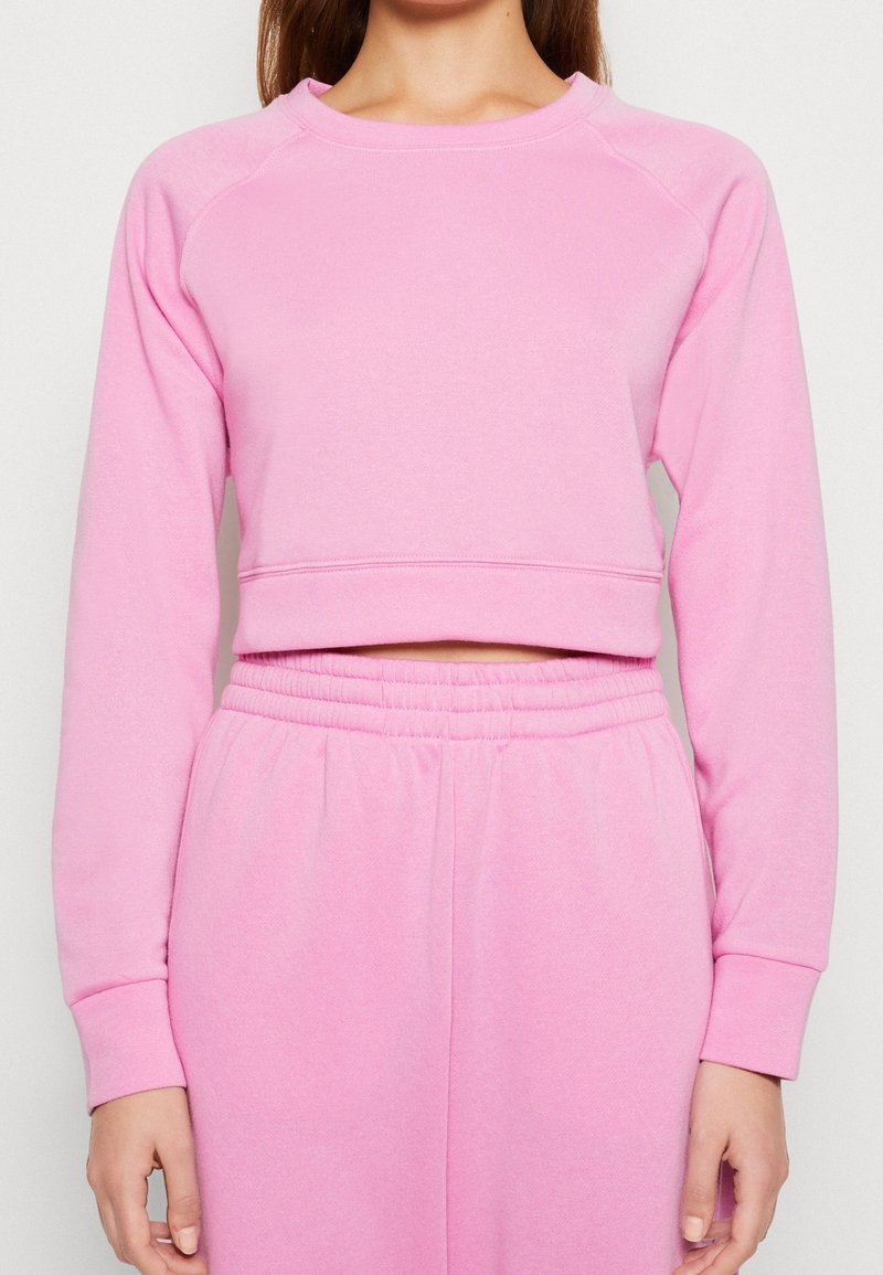 Femme portant un sweat court rose à manches longues et un pantalon de jogging rose taille haute assorti sur un fond uni.