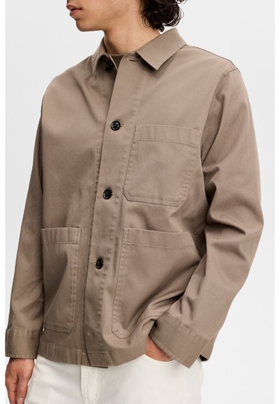 Mężczyzna w taupeowej kurtce na guziki z trzema przednimi kieszeniami i kołnierzem, w białych spodniach, z ręką w kieszeni.