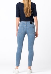 Kvinna med långt hår som bär en marinblå topp, ljusblå skinny jeans med rå kant och vita sneakers, stående vänd bort på vit bakgrund.