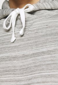 Felpa grigia a righe con una texture morbida, dotata di un laccio bianco e una tasca frontale. Il materiale sembra leggero e informale.