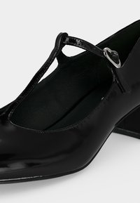 Chaussure Mary Jane noire en cuir verni avec bride en T, petit talon bloc et boucle argentée sur fond clair.
