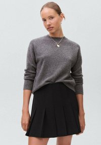 Maglione grigio lavorato a maglia con polsini a costine abbinato a una gonna nera plissettata, caratterizzata da un orlo corto e una texture di tessuto liscia.