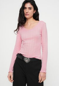 Ροζ ανοιχτόχρωμο τοπ με μακριά μανίκια και ριμπ ύφασμα, με λαιμόκοψη scoop. Συνδυάζεται με μαύρο παντελόνι και ασημένια αγκράφα ζώνης σε σχήμα καρδιάς.