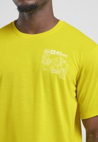 T-shirt di cotone gialla a maniche corte, con una stampa grafica bianca di montagne e il logo "Jack Wolfskin" sul petto.