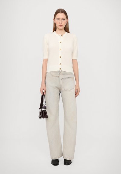 White ribbed cardigan with short sleeves and gold buttons, paired with light wash wide-leg jeans. Holds a small dark handbag, wearing black shoes.