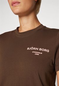 T-shirt in cotone marrone con colletto rotondo, maniche corte e testo stampato di un rosa chiaro: "BJÖRN BORG STOCKHOLM 1956." Texture liscia.