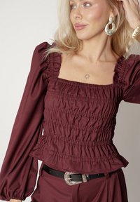 Burgundowy top peplum z marszczonym materiałem, kwadratowym dekoltem i długimi bufkami. Posiada fakturowane wykończenie i subtelny połysk. Czarny pasek z srebnym klamrą.