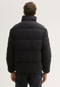 Sort puffjakke med høj krave og quiltet design. Har en glat tekstur og elastiske manchetter, båret over mørke jeans.