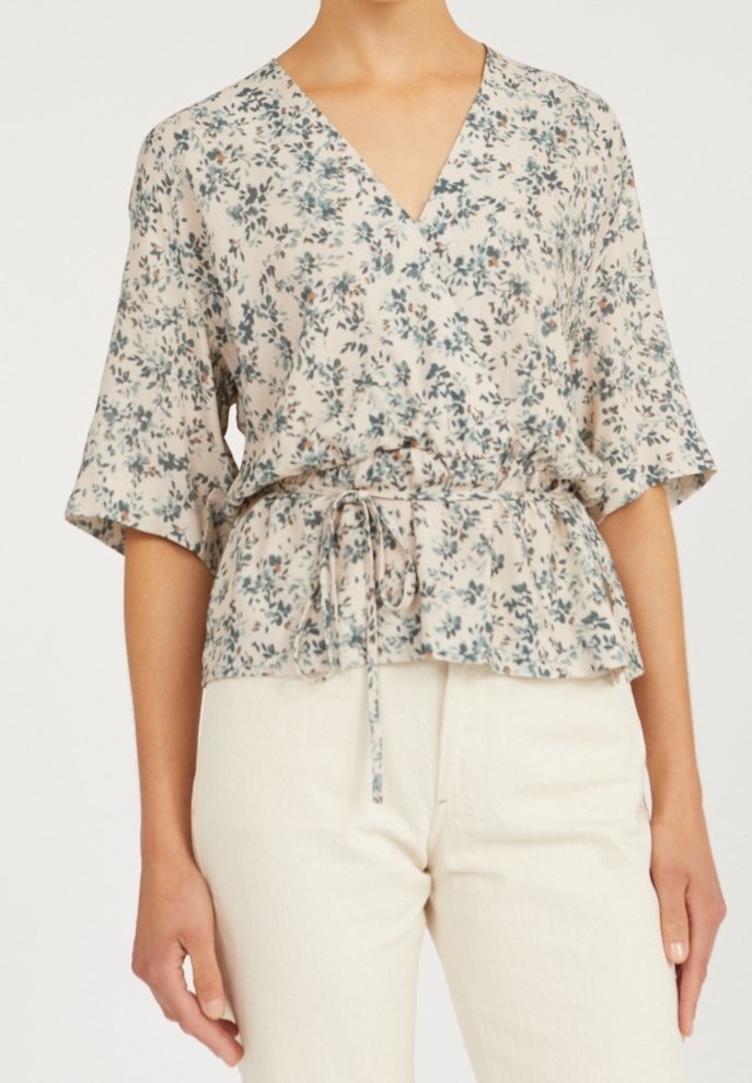 Femme portant un chemisier léger à motif floral avec col en V, manches longueur coude et ceinture à la taille, associé à un pantalon couleur crème.