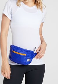 Blå midjeväska gjord av lättviktigt material, med en gul dragkedja och logopatch. Den har en mjuk, texturerad yta och en böjd form.