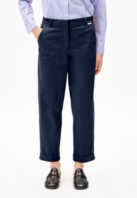 Pantaloni di misto cotone blu navy con taglio dritto, pieghe frontali, tasche laterali e orli arrotolati, indossati con mocassini scuri.