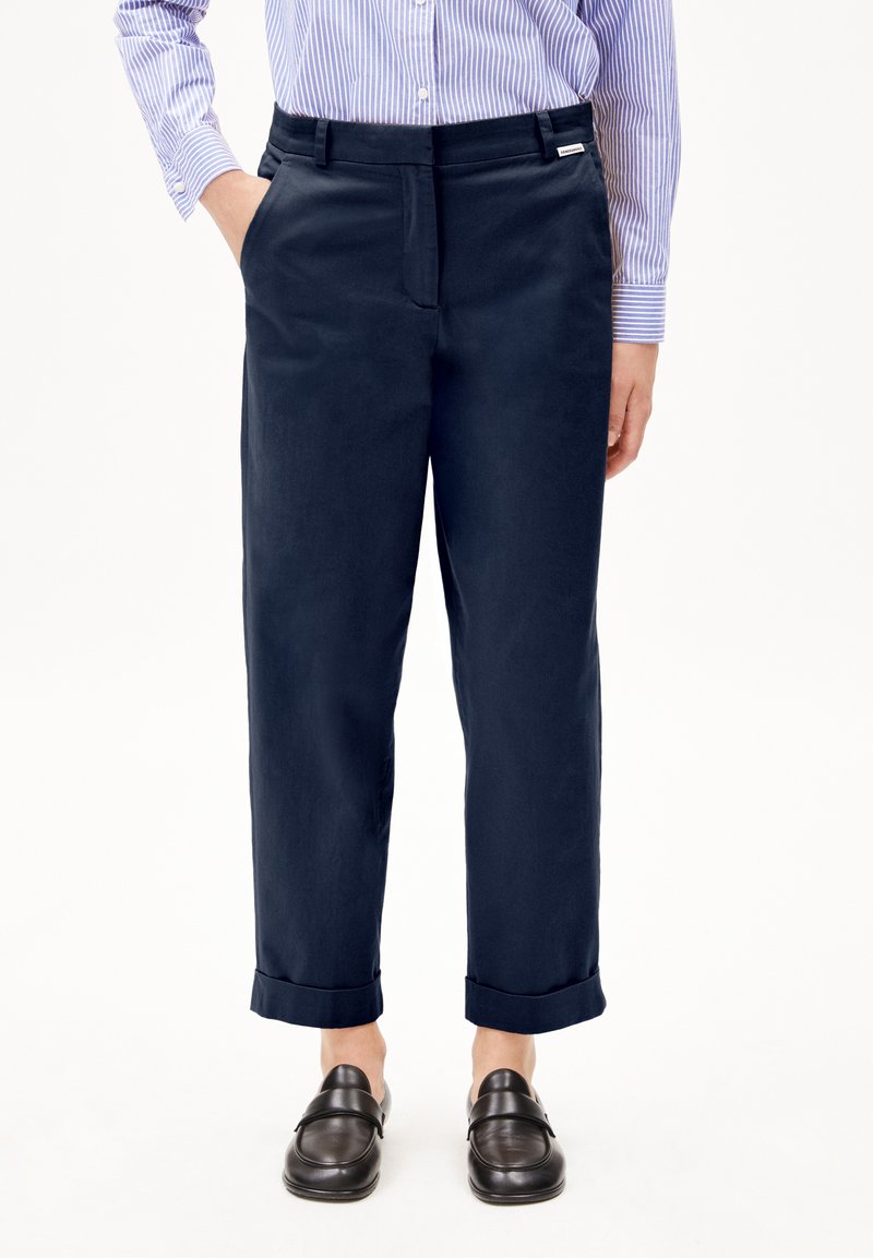 Pantaloni di misto cotone blu navy con taglio dritto, pieghe frontali, tasche laterali e orli arrotolati, indossati con mocassini scuri.