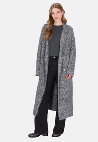 Donna con lunghi capelli mossi, che indossa un lungo cardigan grigio a trame, una maglietta grigia scura, pantaloni neri a vita alta e scarpe nere, in piedi.