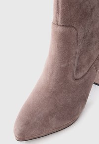 Taupe suede ankle boot featuring a pointed toe and a curved seam detail. The stacked heel adds height and stability to the design.