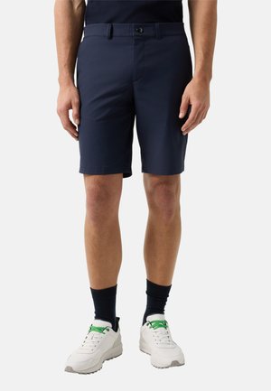 Mann trägt navyblaue, maßgeschneiderte Shorts, schwarze Socken und weiße Turnschuhe mit grünen Schnürsenkeln, steht vor einem schlichten Hintergrund.