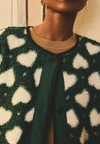 Giacca verde con texture e motivi a cuore bianchi, con un bordino nero a contrasto e una chiusura con bottoni in evidenza. Sotto, un maglione beige.