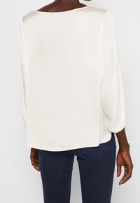 Weiße Satinbluse mit rundem Ausschnitt, schwingenden Ärmeln und einem leicht lockeren Schnitt, kombiniert mit dunkelblauen Jeans.