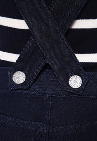 Les salopettes en denim présentent une couleur bleu foncé, des bretelles croisées et un bouton métallique avec un logo. Des rayures blanches horizontales sont visibles sur le tissu.