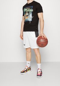 Camiseta gráfica negra con estampado colorido, pantalones cortos deportivos blancos con acentos negros y zapatillas deportivas multicolores. Sosteniendo un balón de baloncesto.