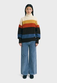 Gebreide oversized trui met een hoge hals, voorzien van horizontale strepen in crèmekleurig, geel, oranje, donkerbruin en blauw; gecombineerd met brede blauwe corduroy broek.