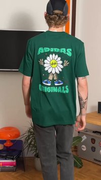 Žalia medvilninė marškinėliai su dideliu baltu ramunės grafiniu vaizdu, geltonu centru ir užrašu „ADIDAS ORIGINALS“. Modelis dėvi pilkas kelnes.