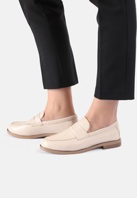 Béžové kožené mokasíny s plochou podr�ážkou a prošitím, nošené s černými dobře padnoucími kalhotami, které ukazují elegantní a minimalistický design.