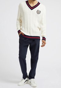 Homme portant un pull blanc à col en V tricoté en câble avec des bordures rouges et bleu marine, un pantalon bleu marine foncé et des baskets blanches, debout avec une main dans la poche.