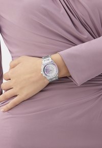 Naise käsi toetub puusale, käELs hõbedane metallkell, millel on roosa ja lilla ehetega dial ja käevõru, seljas mauve värvi volditud kleit.