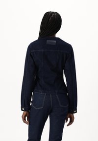 Giacca in denim scuro con una vestibilità su misura, maniche lunghe e un'etichetta in pelle sul retro. Dettagli con cuciture a contrasto e polsini con bottoni.