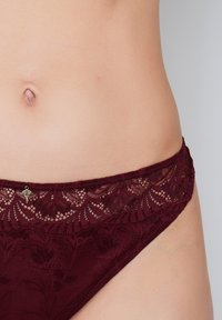 Burgundy lace bikini bottom with floral pattern, featuring a scalloped edge and small metallic charm on the waistband. Soft texture.