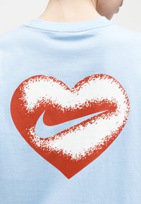 Άτομο που φοράει ανοιχτό μπλε πουκάμισο με ένα μεγάλο κόκκινο και λευκό γραφικό σε σχήμα καρδιάς, στο οποίο υπάρχει το λογότυπο Nike σε λευκό, στην πλάτη.