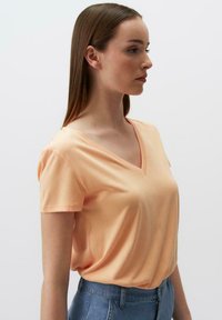 T-shirt con scollo a V in pesche realizzata in morbido cotone, con maniche corte, vestibilità rilassata e texture liscia, abbinata a pantaloni denim a vita alta.