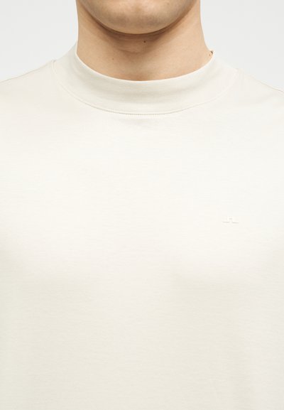 Κοντινό πλάνο ενός ατόμου που φοράει μια απλή ανοιχτόμπεζ μπλούζα με ψηλό λαιμό και ένα μικρό, διακριτικό λογότυπο στο στήθος.