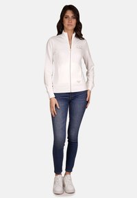 Giacca bianca con zip, dal taglio slim fit, colletto alto e dettagli con logo discreto. Abbinata a jeans scuri e sneaker bianche, mostrando un outfit informale.
