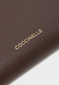 Sötétbarna bőrtárca, amelynek textúrázott felülete van, és arany "Coccinelle" márkázás díszíti. Kompakt kialakítás, látható varrási részletekkel.