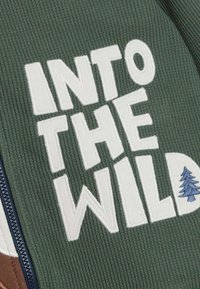 Zaļš audums ar ribotu tekstūru, uz kura ir izšūtas drosmīgas baltas burti "INTO THE WILD" un neliela zila koka grafika. Zipsera redzama sānos.