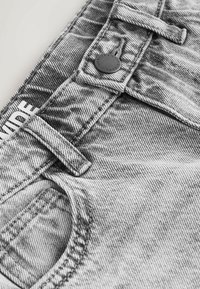 Valge tekstiga valge sildi osaliselt nähtavusega helehallide teksapükste lähivõte, kus on nähtavad nööp, vööaasad ja esikotitipp.