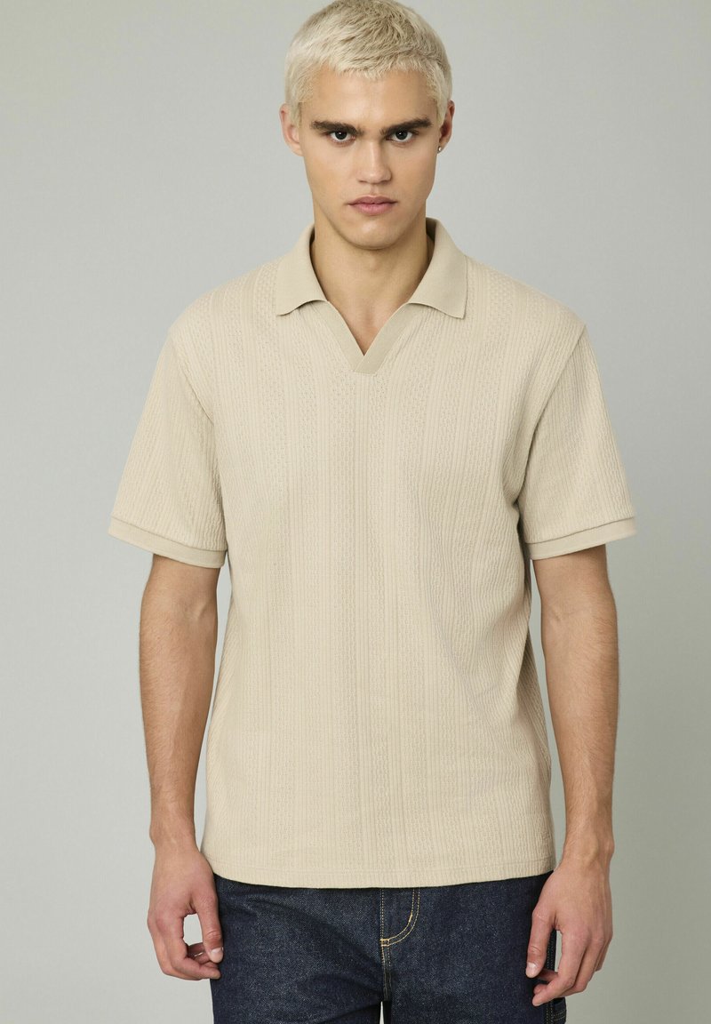 Polo en maille beige avec un col en V, des manches courtes et une texture côtelée. Conçu avec une coupe décontractée et de légères rayures verticales.