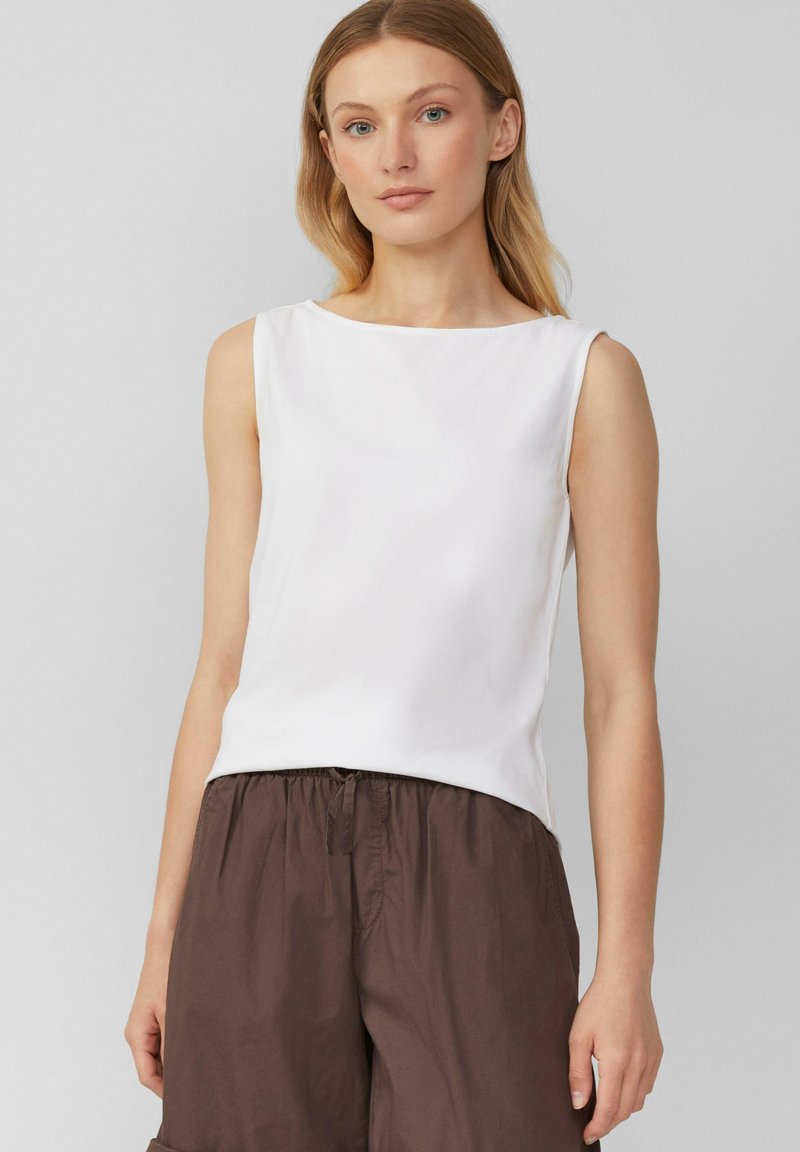 Top blanco sin mangas con escote barco, hecho de una tela suave. Combinado con pantalones cortos marrones holgados con cintura elástica.