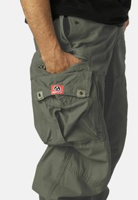Olive zelene cargo hlače z velikim stranskim žepom, majhno rdečo nalepko z logotipom in več podvojenimi šivi.