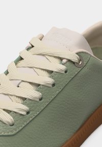 Gros plan sur une sneaker en cuir texturé vert avec des lacets crème, une languette blanche et une semelle en caoutchouc marron.