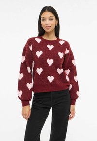 Bordo džemper s uzorkom ruži�častih srca, dugim rukavima, okruglim izrezom i mekanom, teksturiranom završnom obradom. Uparen s crnim hlačama.