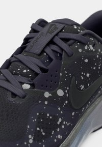 Zapatilla atlética negra y gris oscuro con patrón de salpicaduras, que presenta una parte superior texturizada, cordones planos y una suela de goma con un acento translúcido.