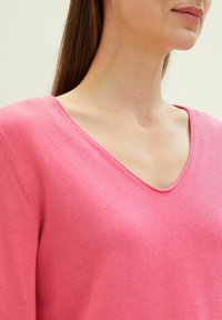 Rosa gestrickter Pullover mit V-Ausschnitt. Die Textur wirkt glatt mit dezenten Nahtdetails entlang des Halsausschnitts.