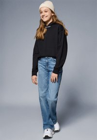 Svart langermet genser med Calvin Klein-logo, lyseblå denimjeans og hvite sneakers. Modellen har på seg en beige ribbestrikket lue.