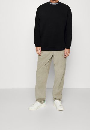 Felpa nera con il testo "BROOKLYN" sul colletto, abbinata a pantaloni di velluto a coste beige e sneakers bianche con dettagli verdi.