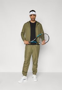 Oliven grønn treningsjakke med glidelås og tettsittende bukser, svart t-skjorte underneath, hvit visir og en tennisracket. Hvite joggesko.