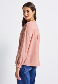 Langärmlige, pinke Bluse mit rundem Ausschnitt und gerafften Manschetten, kombiniert mit blauen Jeans. Der Stoff wirkt weich und fließend.