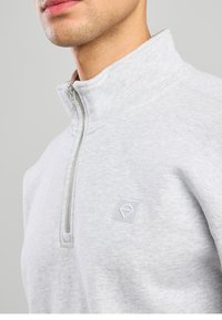 Gray zip-up sweatshirt made of soft fabric, featuring a high collar and a logo patch on the chest with a textured pattern.